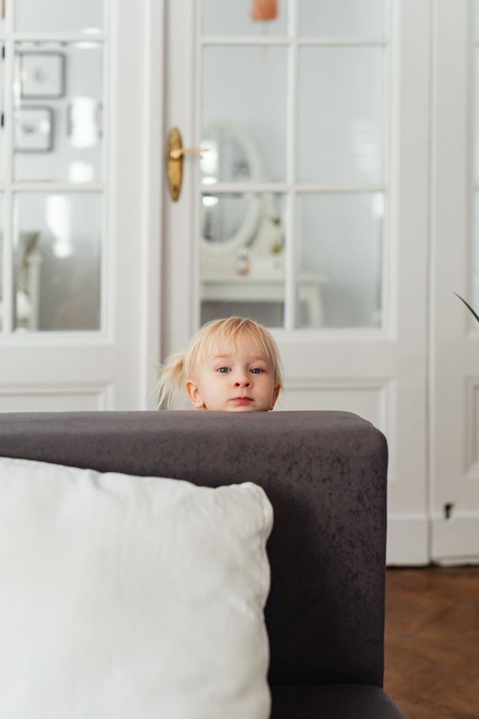 About A curious blonde child peeks over the couch in a cozy room with elegant decor and glass doors.